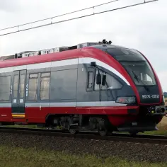 Czerwono-szary pociąg elektryczny ED74-001d na torach, w tle pole i niebo.